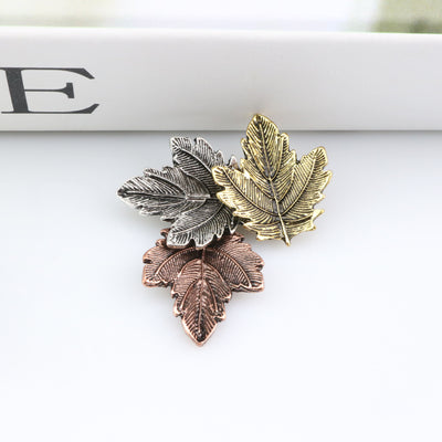 Three leaf-shaped pins in gold, silver, and bronze on a white surface.