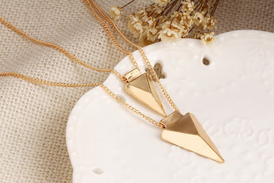 Gold necklaces with geometric pendants on a textured surface with dried flowers.