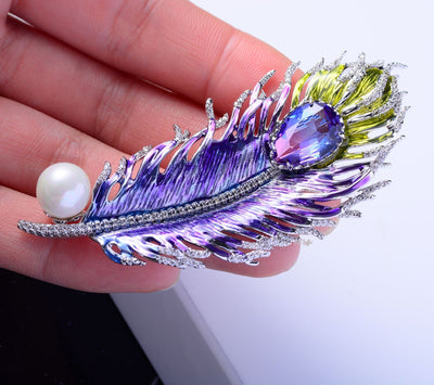 Decorative feather brooch with gemstones and pearl held between fingers against a white background