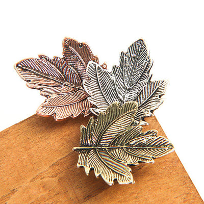 Three leaf-shaped pins in bronze, silver, and gold on a wooden surface.