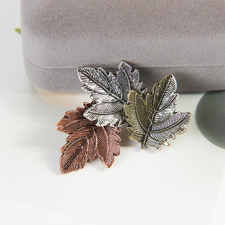 Two leaf-shaped brooches in silver and bronze on a white surface with a gray box in the background.