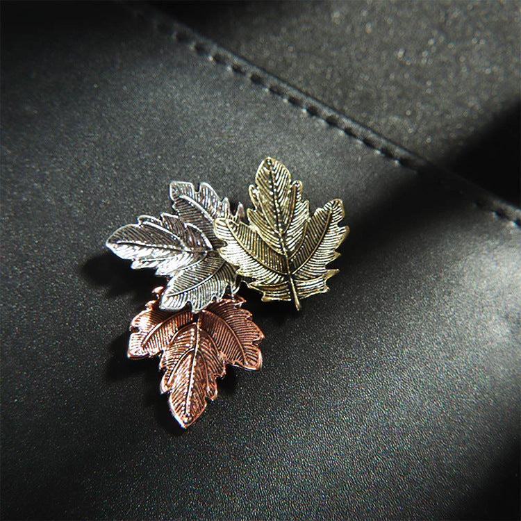 Three leaf-shaped pins in silver, gold, and rose gold on a dark surface