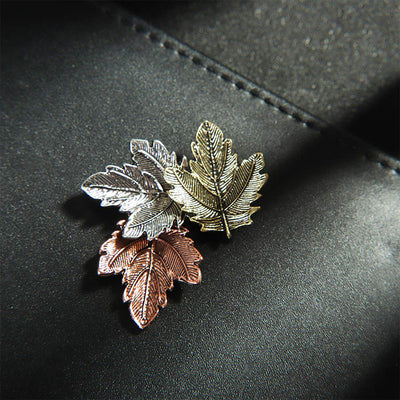 Three leaf-shaped pins in silver, gold, and rose gold on a dark surface