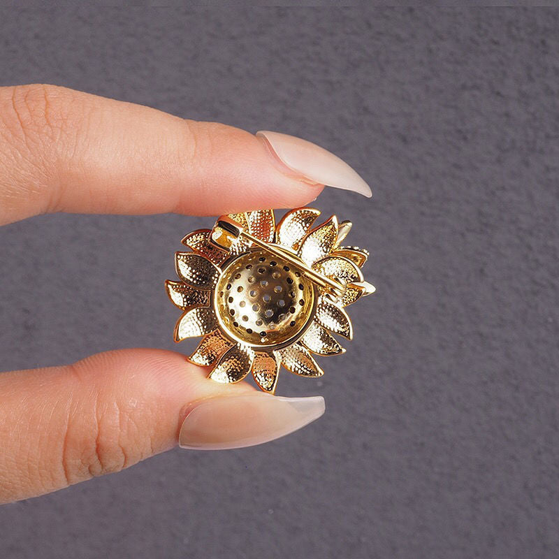 the back of a Gold sunflower-shaped brooch held between fingers against a gray background