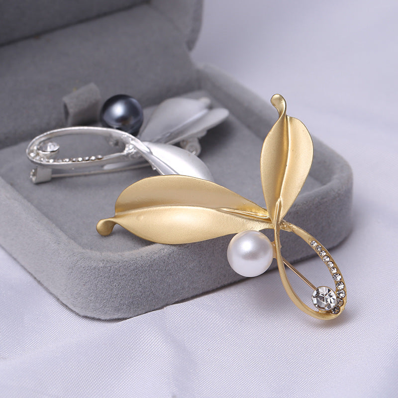 silver and a Gold leaf-shaped brooch with pearl and gemstone on a gray jewelry box