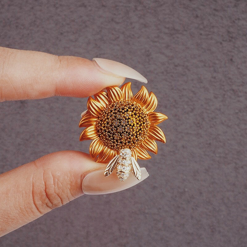 Gold sunflower-shaped earring with a bee detail held between fingers against a gray background