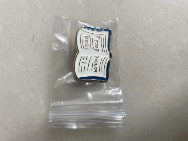 Pin shaped like an open book with text in a clear plastic bag on a gray background
