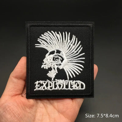 Black patch with white embroidered design and 'Exploited' text held in a hand against a dark background.