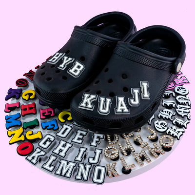 Black clogs with customizable letter charms on a pink background