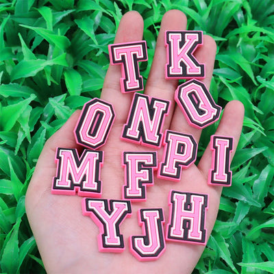 Hand holding pink and black letter shoe charms against a green grass background