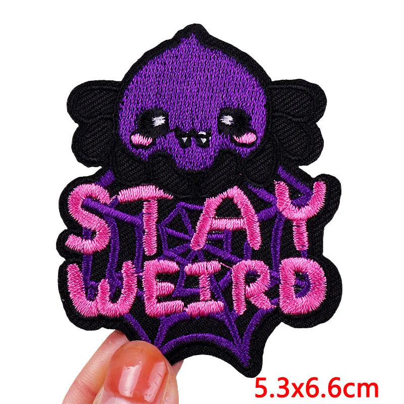 Embroidered patch with a purple skull and 'Stay Weird' text held by a hand.