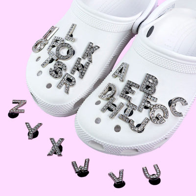 White clogs with shoe charms rhinestone letter charms on a pink background