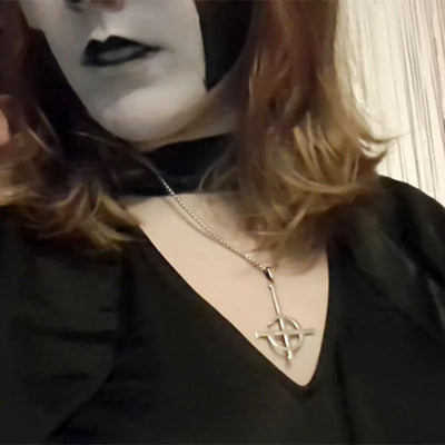 Person wearing a black outfit with a necklace featuring a symbol, against a neutral background.