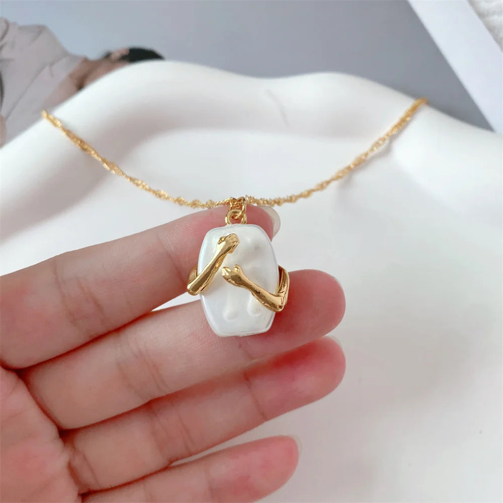 A hand holding a Gold necklace with Pendant of 2 hands hugging a white Natural iridescent stone pendant against a  white Dish