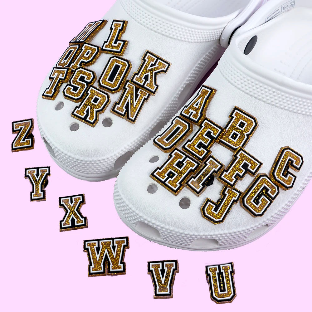 White clogs with gold shoe charms lettering on a pink background