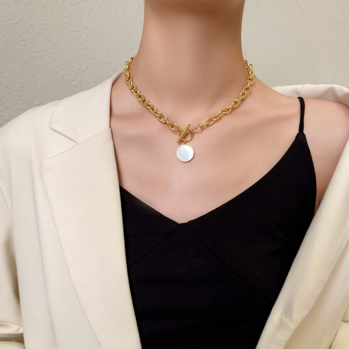 Gold chain necklace with a round pendant worn by a person in a black top and white blazer.