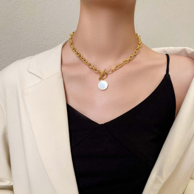 Gold chain necklace with a round pendant worn by a person in a black top and white blazer.