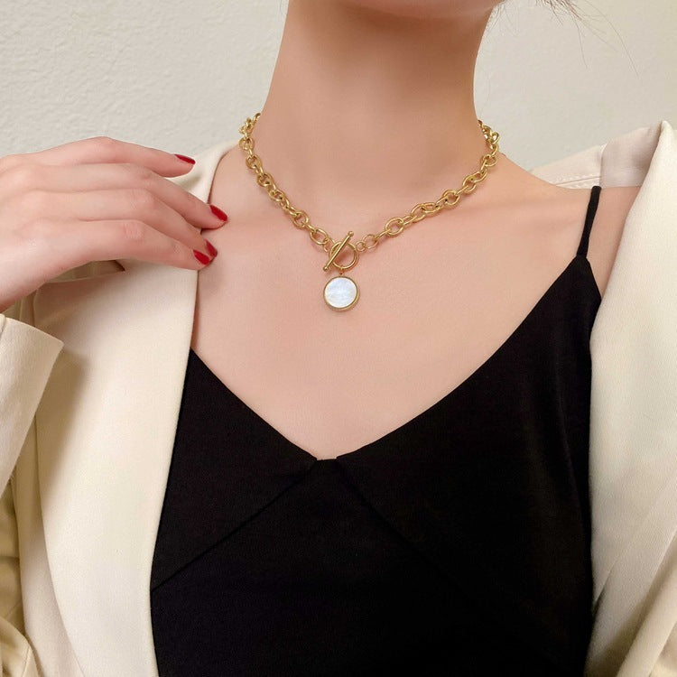 Person wearing a gold necklace with a round pendant, dressed in a black top and beige jacket.