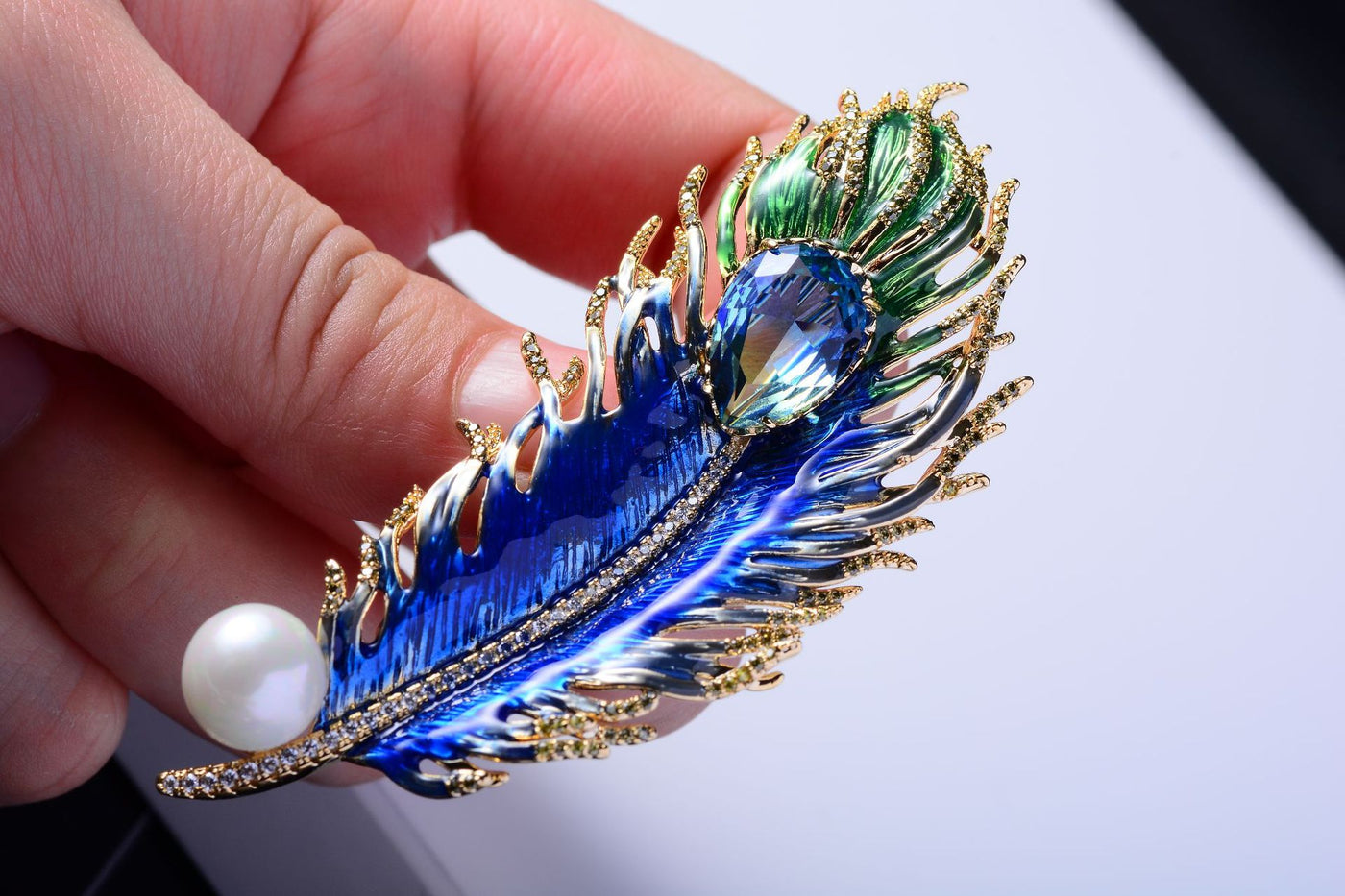 Decorative brooch with peacock feather design held by a hand against a light background